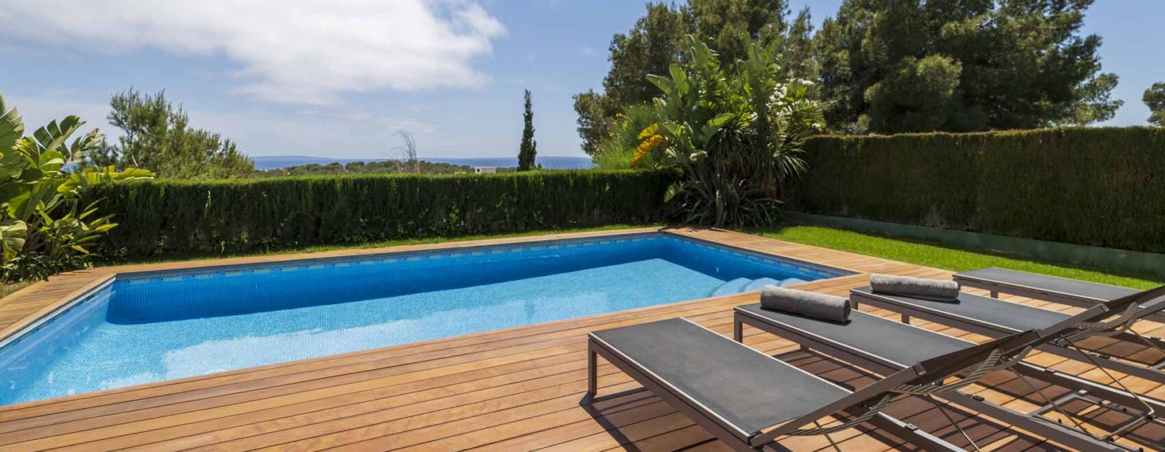Luxurious private pool with crystal blue water, flanked by a spacious wooden deck and comfortable sun loungers, with lush greenery and a view of the sea horizon in the background.