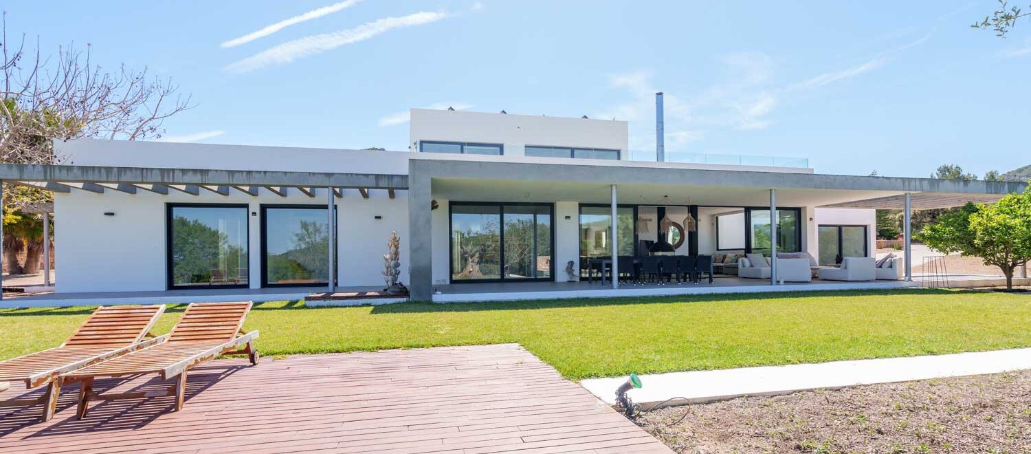 Moderna villa blanca en venta con gran piscina y terraza de madera en Benimussa, Ibiza, con un exuberante césped verde y un cielo azul despejado.