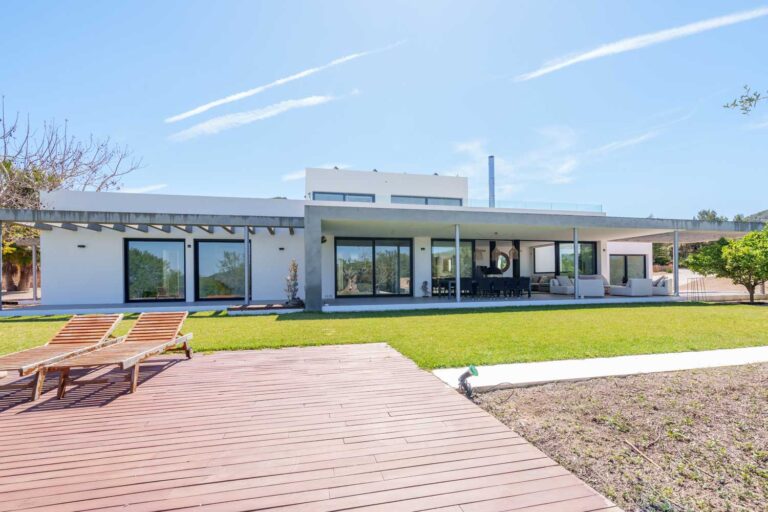 Modern white villa for sale with large swimming pool and wooden deck in Benimussa, Ibiza, showcasing lush green lawn and clear blue skies