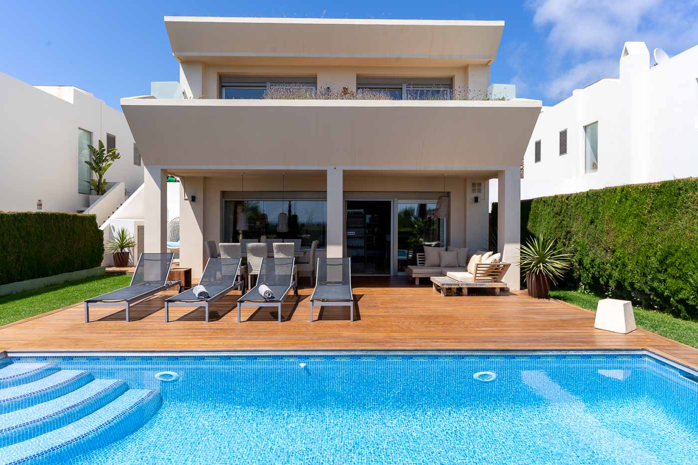 Modern white villa with expansive sliding doors opening onto a sun-drenched deck with stylish outdoor furniture, leading to a stunning pool with steps, all set against a backdrop of manicured hedges and clear blue skies.
